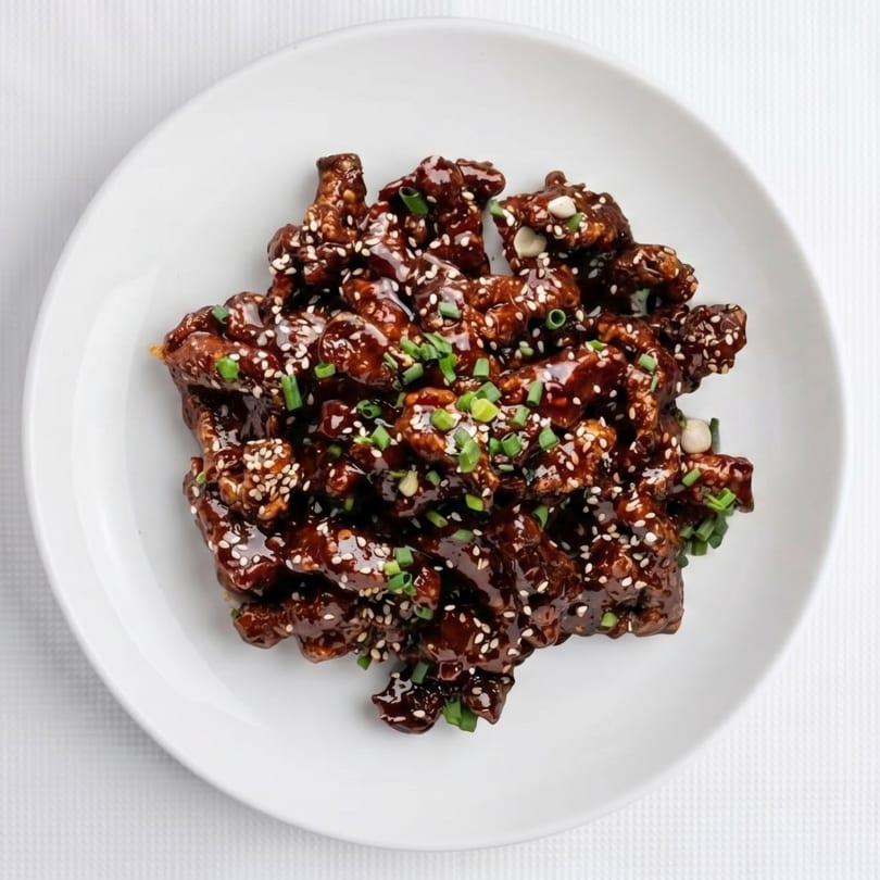 Sweet and sour chicken, garnished with sesame seeds and chopped green onions, on a white plate.