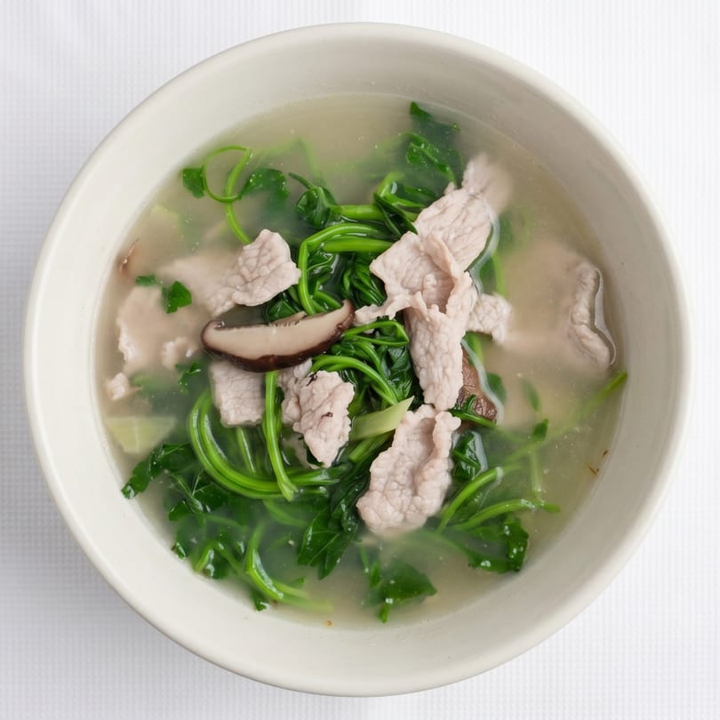 Overhead view of a clear soup with green leafy vegetables, sliced pork, and mushrooms in a white bowl.