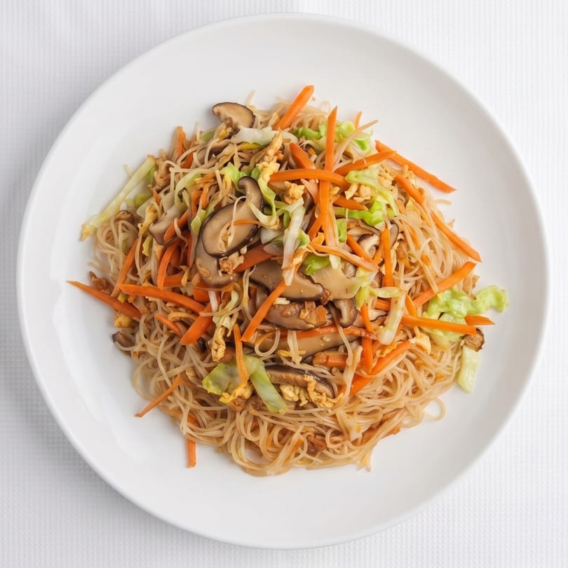 A white plate holds a serving of stir-fried rice vermicelli noodles with vegetables and mushrooms.